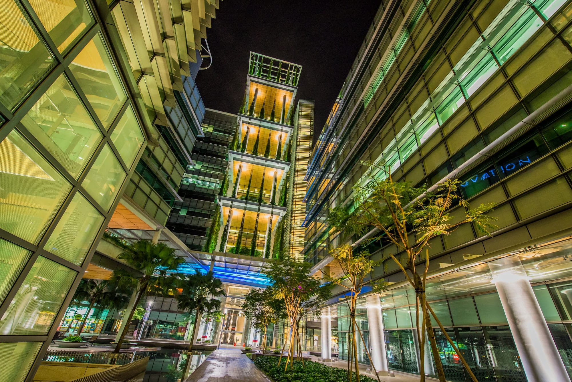 Glass towers at night framing a warm-lit tower with uplit hanging greenery above a landscaped walkway